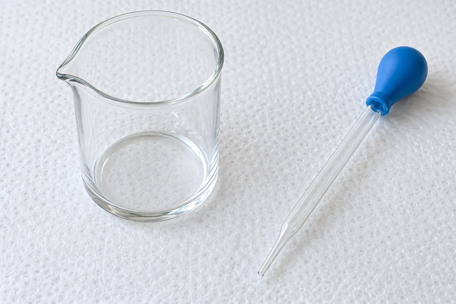 Un recipiente de vidrio sin marcas y una pipeta transparente con pera azul descansan sobre una superficie blanca